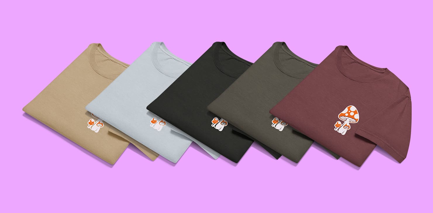 Fünf ordentlich gefaltete T-Shirts in den Farben Beige, Hellgrau, Schwarz, Olivgrün und Dunkelrot liegen nebeneinander auf einem pinken Hintergrund. Auf der Brust jedes Shirts ist ein kleines Motiv mit einem rot-weißen Fliegenpilz und zwei kleineren Pilzen zu sehen.