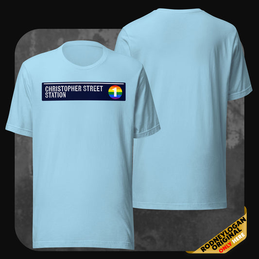 A sky-blue t-shirt featuring the “Christopher Street Station” design and a circular rainbow subway icon on the chest; back of shirt is blank.